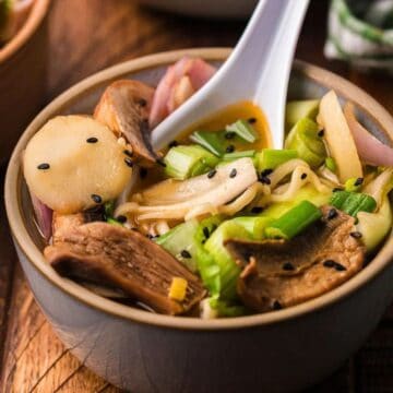 Bowl of ramen with mushrooms, green onions, sliced vegetables, and black sesame seeds, with a white spoon.
