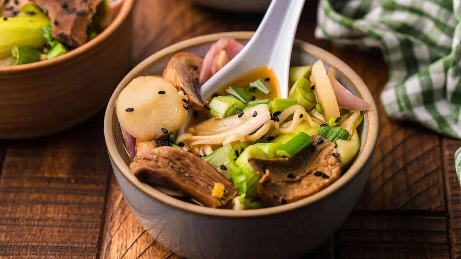 Bowl of ramen with mushrooms, green onions, sliced vegetables, and black sesame seeds, with a white spoon.