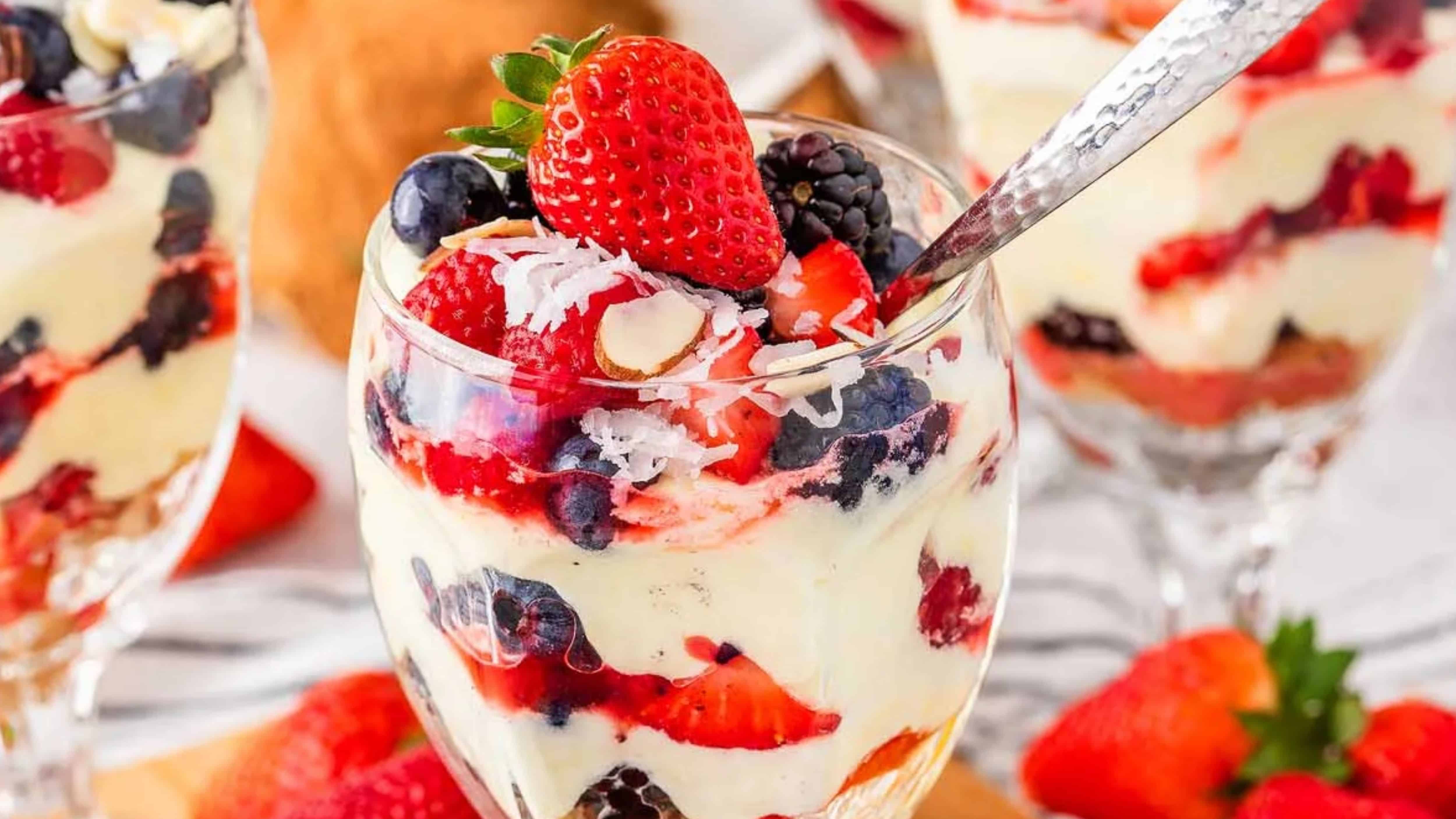 A parfait glass filled with creamy dessert, fresh berries, coconut flakes, and almonds, topped with a whole strawberry.