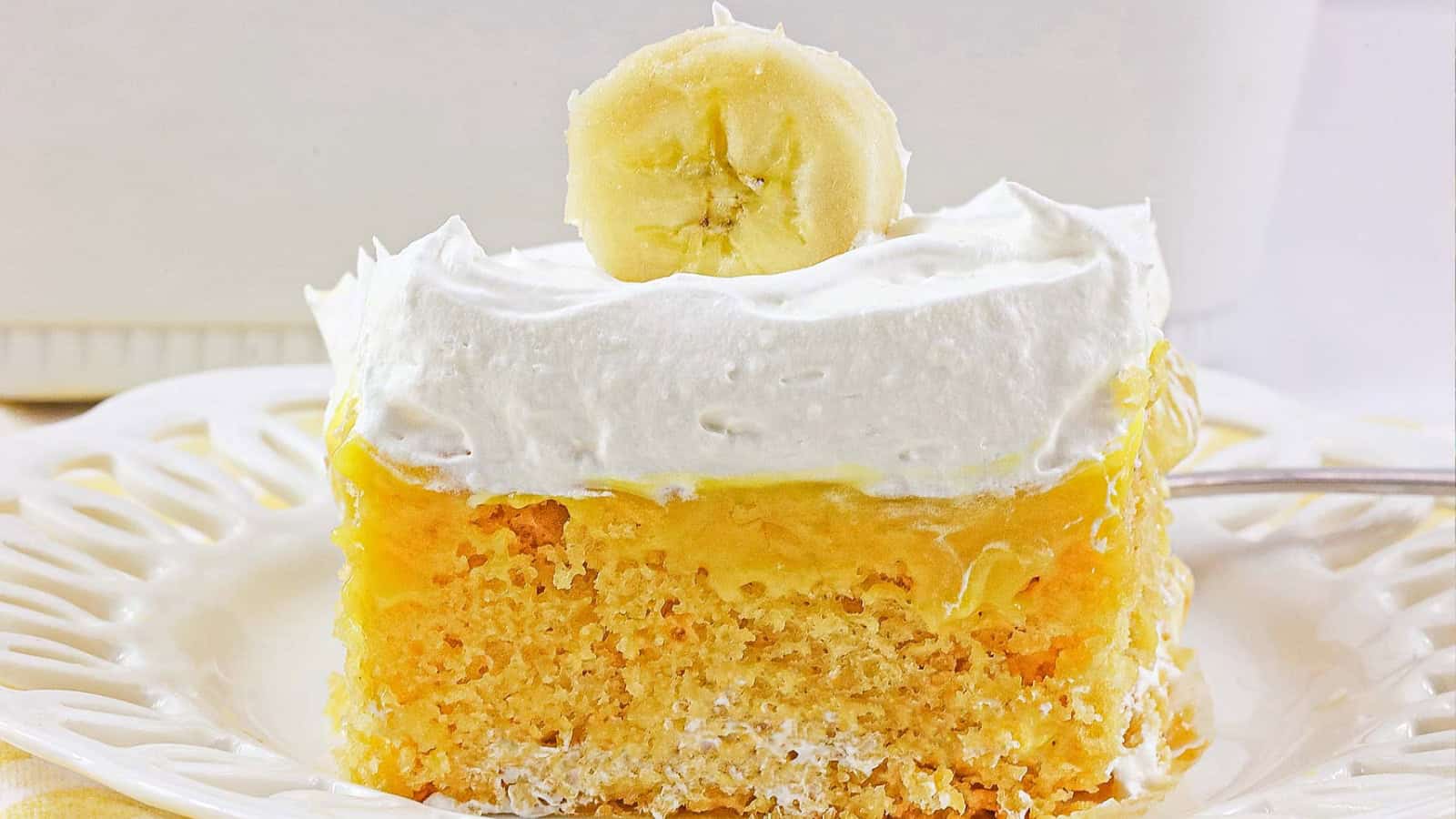A slice of banana pudding cake topped with whipped cream and a banana slice on a white plate.