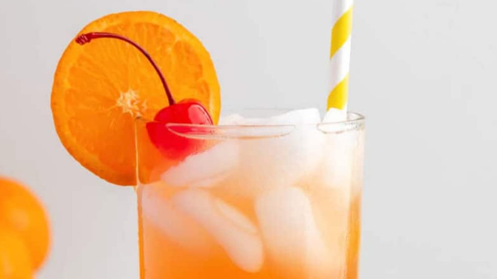 A glass of orange drink with ice, garnished with an orange slice, a cherry, and a yellow-striped straw.