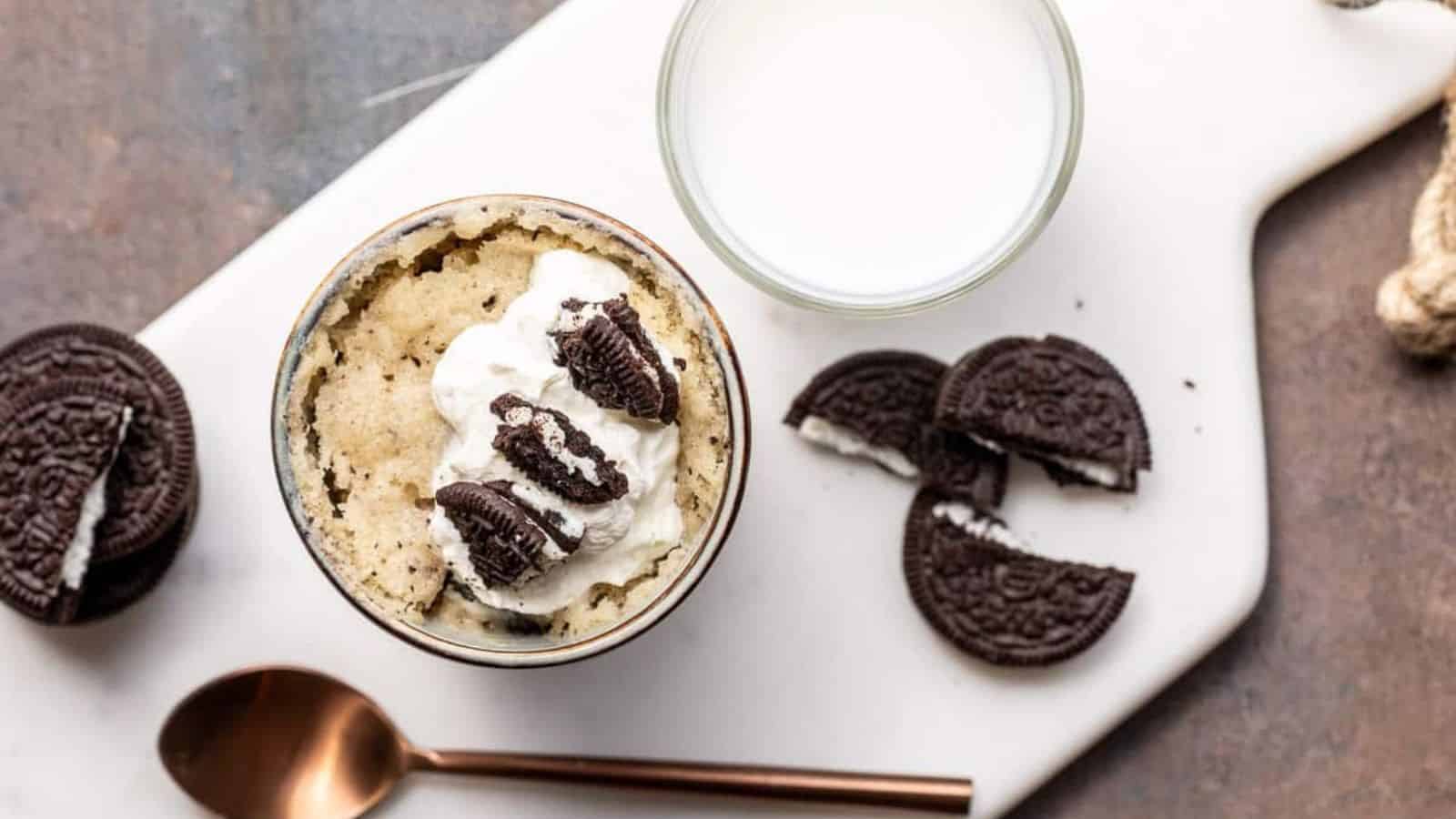 A mug cake with whipped cream and cookie pieces next to a glass of milk and chocolate sandwich cookies.