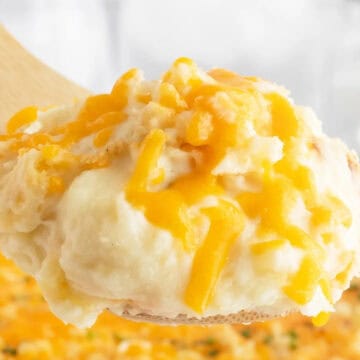 A wooden spoon holds cheesy mashed potatoes topped with melted cheddar cheese, with a casserole in the background.