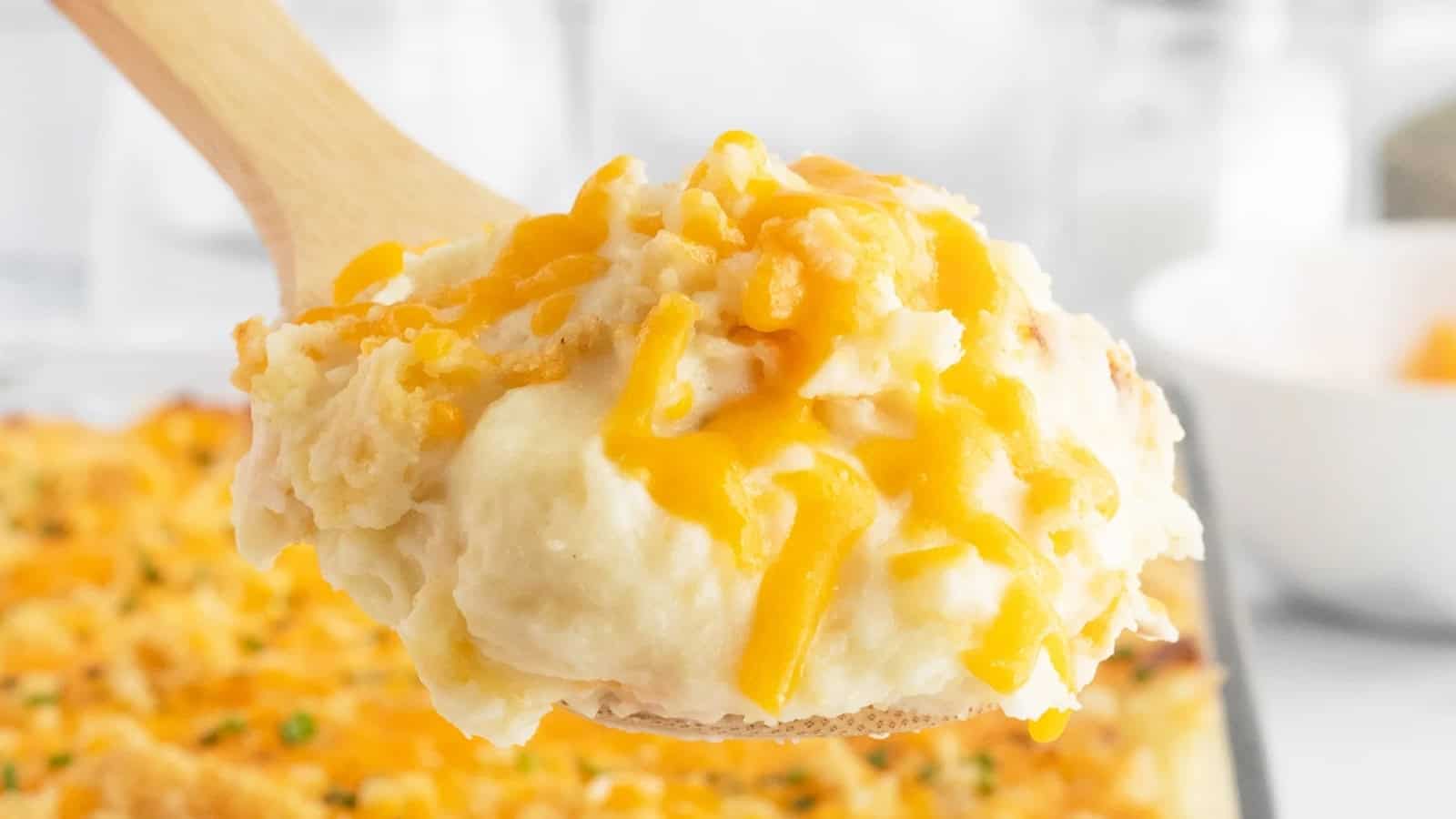 A wooden spoon holds cheesy mashed potatoes topped with melted cheddar cheese, with a casserole in the background.