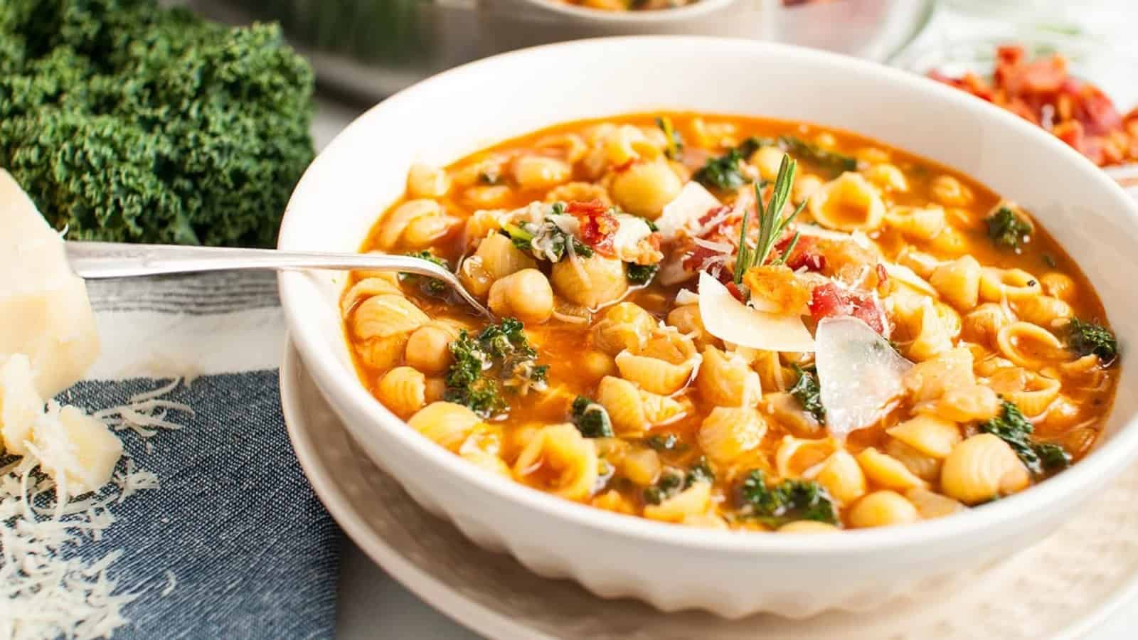 Bowl of pasta soup with kale, chickpeas, tomato broth, herbs, and parmesan cheese on top.