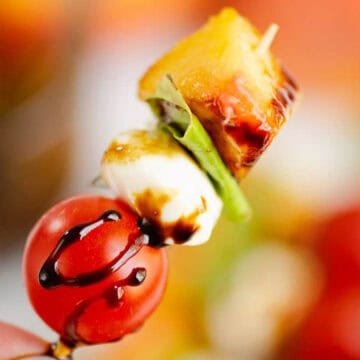 A hand holds a skewer with tomato, mozzarella, basil, pineapple, and balsamic drizzle.
