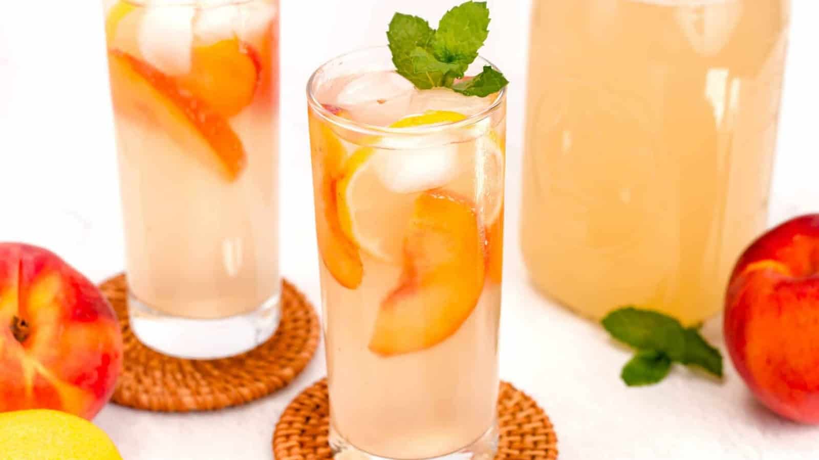 Tall glasses of iced peach lemonade with peach slices and mint on coasters, surrounded by fresh peaches.