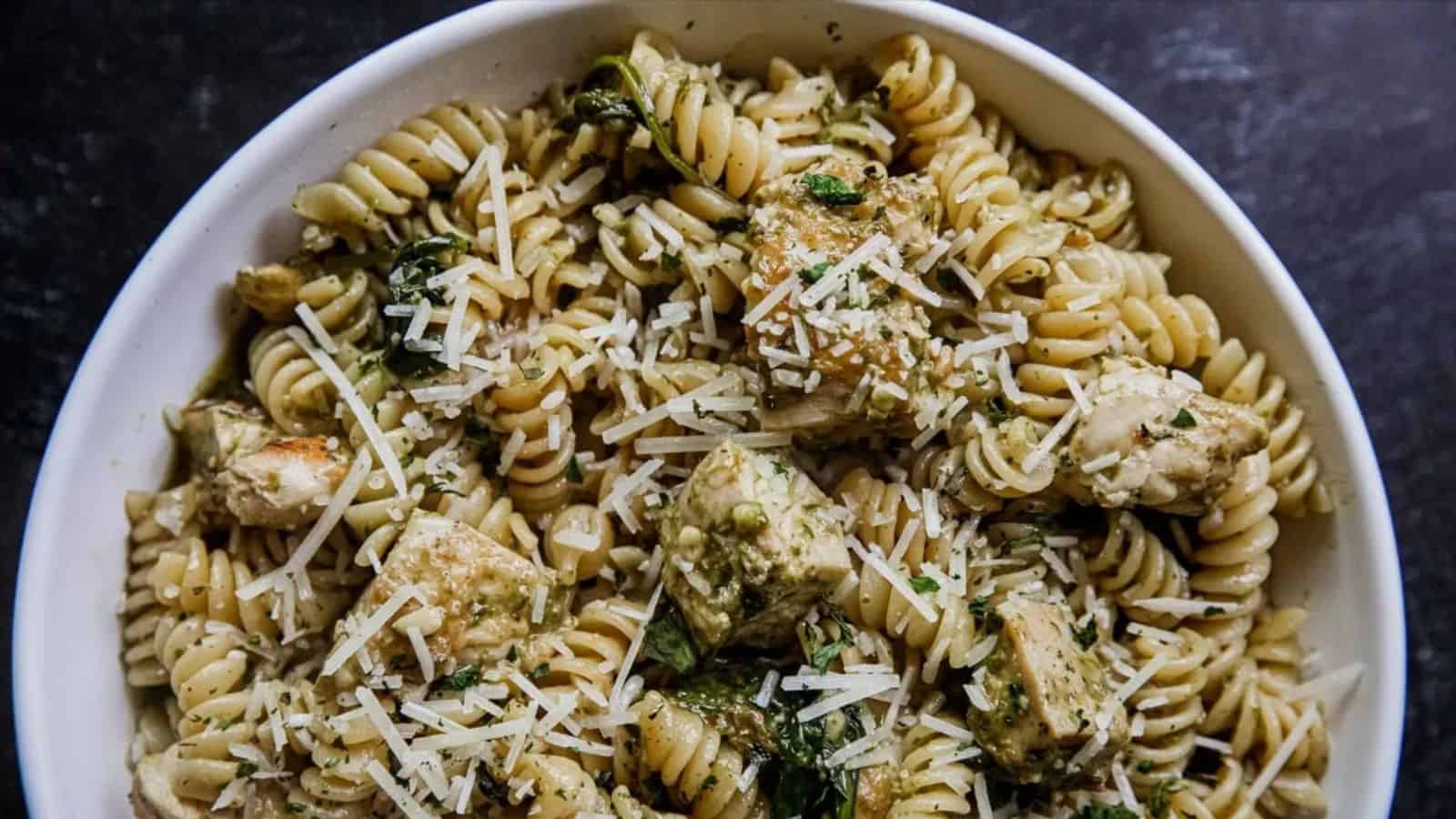 A bowl of rotini pasta with grilled chicken, spinach, pesto sauce, and shredded parmesan cheese.