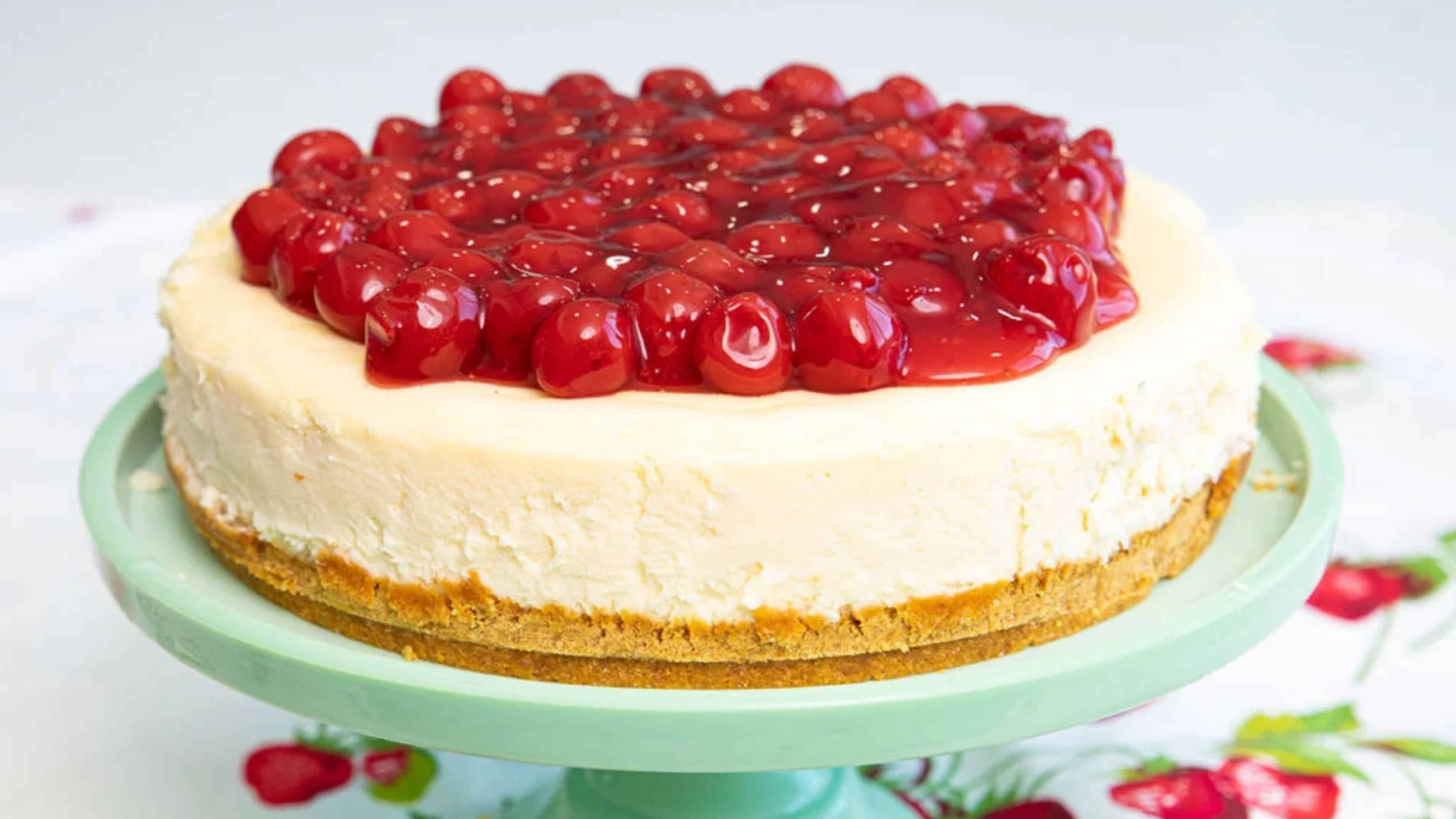 A cheesecake topped with glazed cherries sits on a light green cake stand.