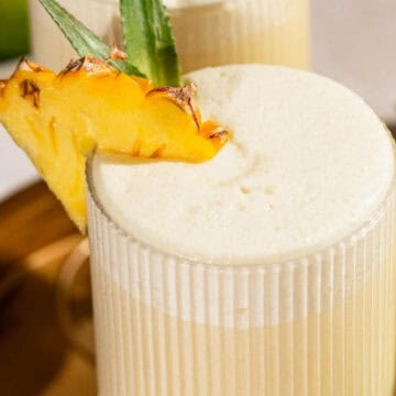 A creamy piña colada garnished with a pineapple slice in a ribbed glass, served on a golden tray.