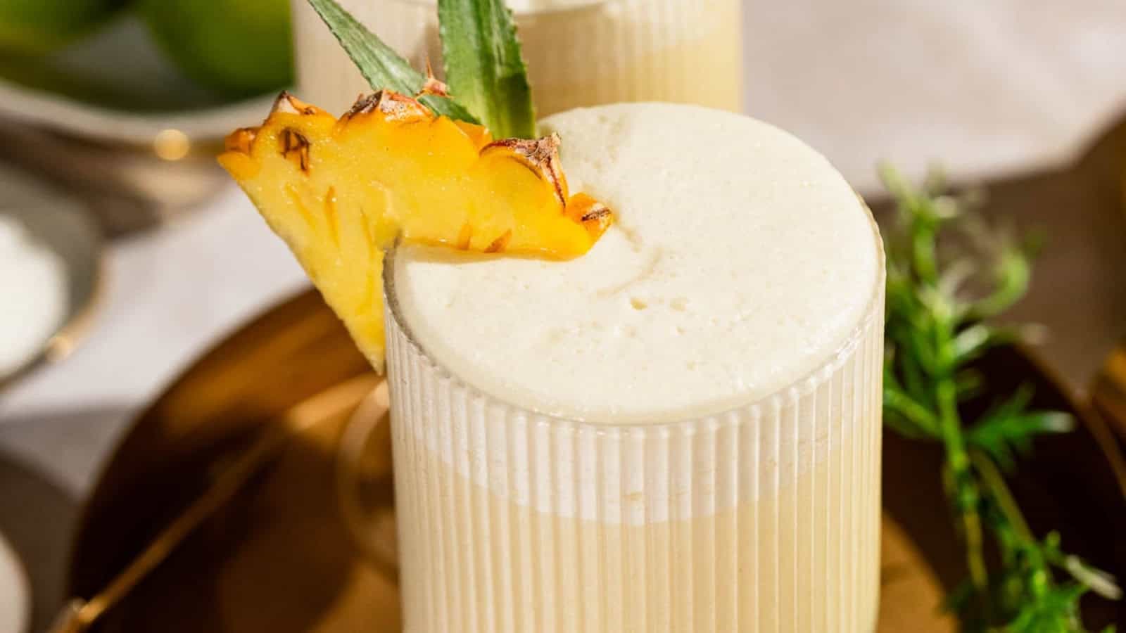 A creamy pi&ntilde;a colada garnished with a pineapple slice in a ribbed glass, served on a golden tray.