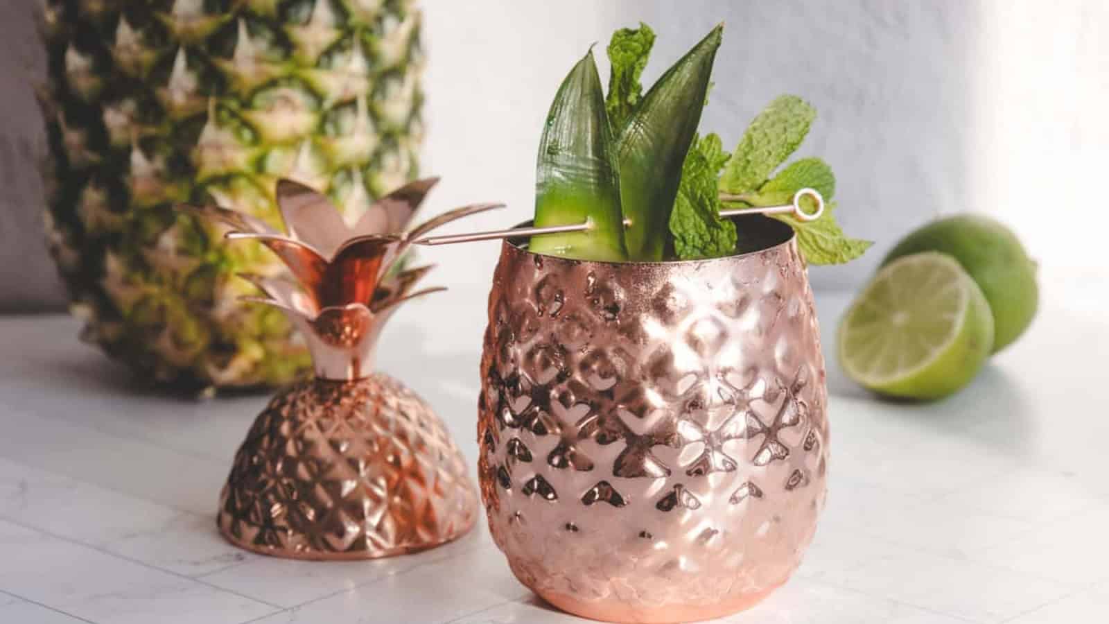 Copper pineapple cup with a leafy cocktail garnish, next to a pineapple and sliced limes on a white surface.