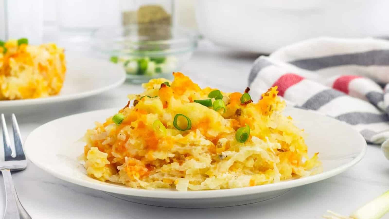 A plate of cheesy hashbrown casserole topped with chopped green onions, with a fork and striped napkin nearby.