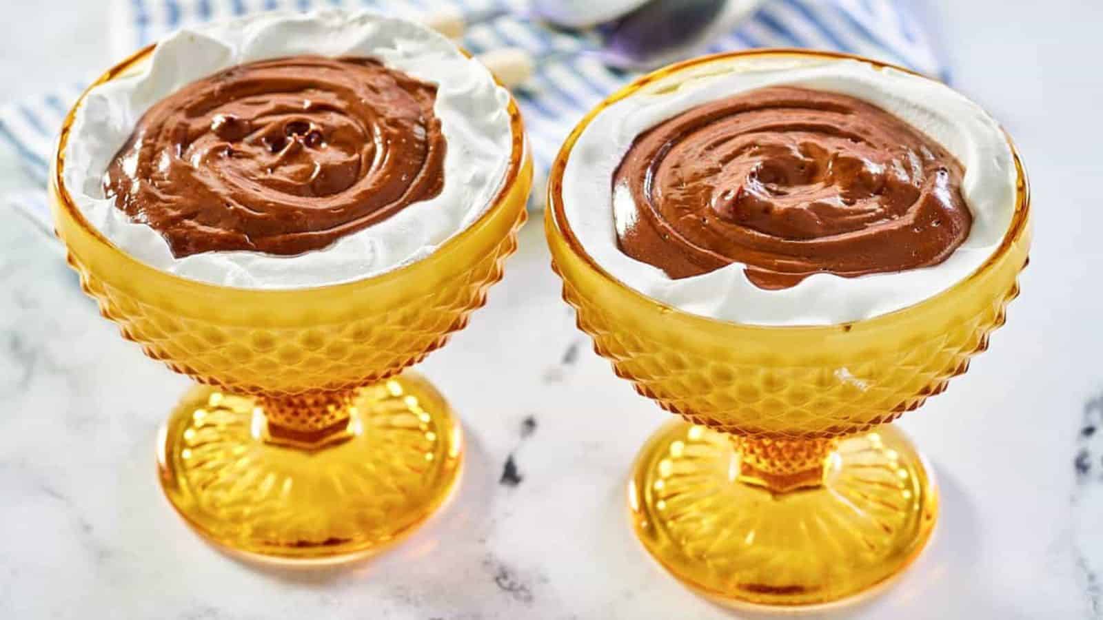 Two yellow glass dessert cups filled with chocolate pudding topped with whipped cream on a marble surface.