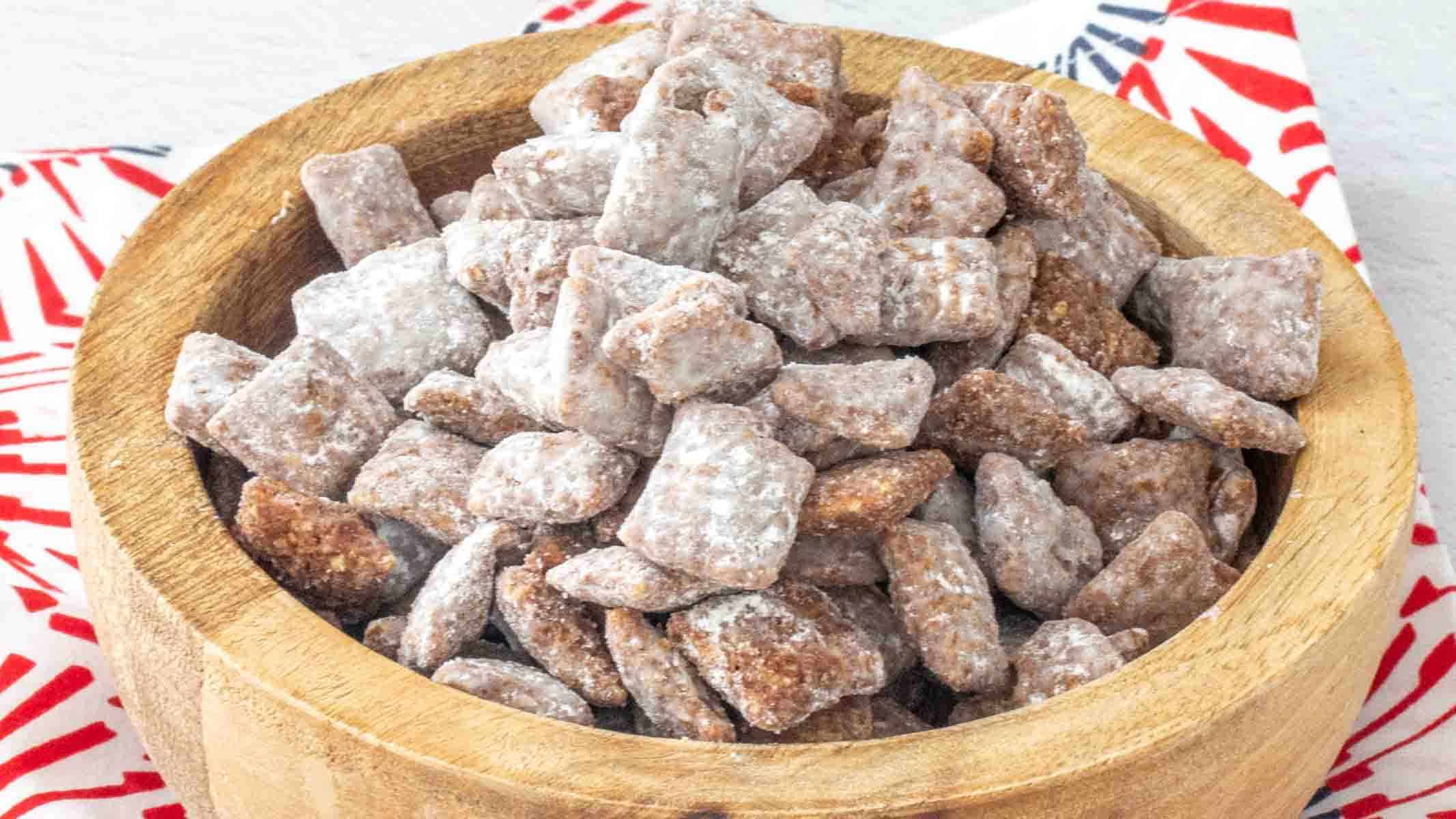 A wooden bowl filled with chocolate-coated, powdered sugar snack mix, often called