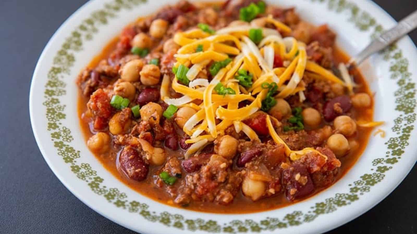 A bowl of chili with chickpeas, beans, cheese, chopped green onions, and ground meat.