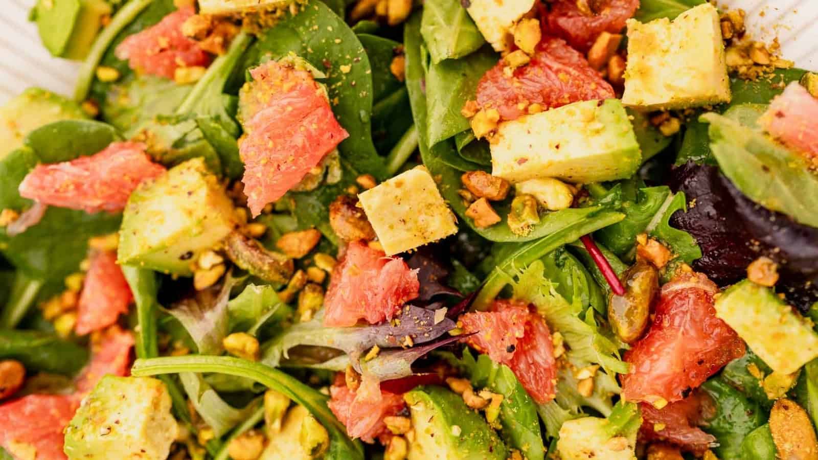 Close-up of a salad with leafy greens, avocado cubes, grapefruit pieces, and chopped nuts.
