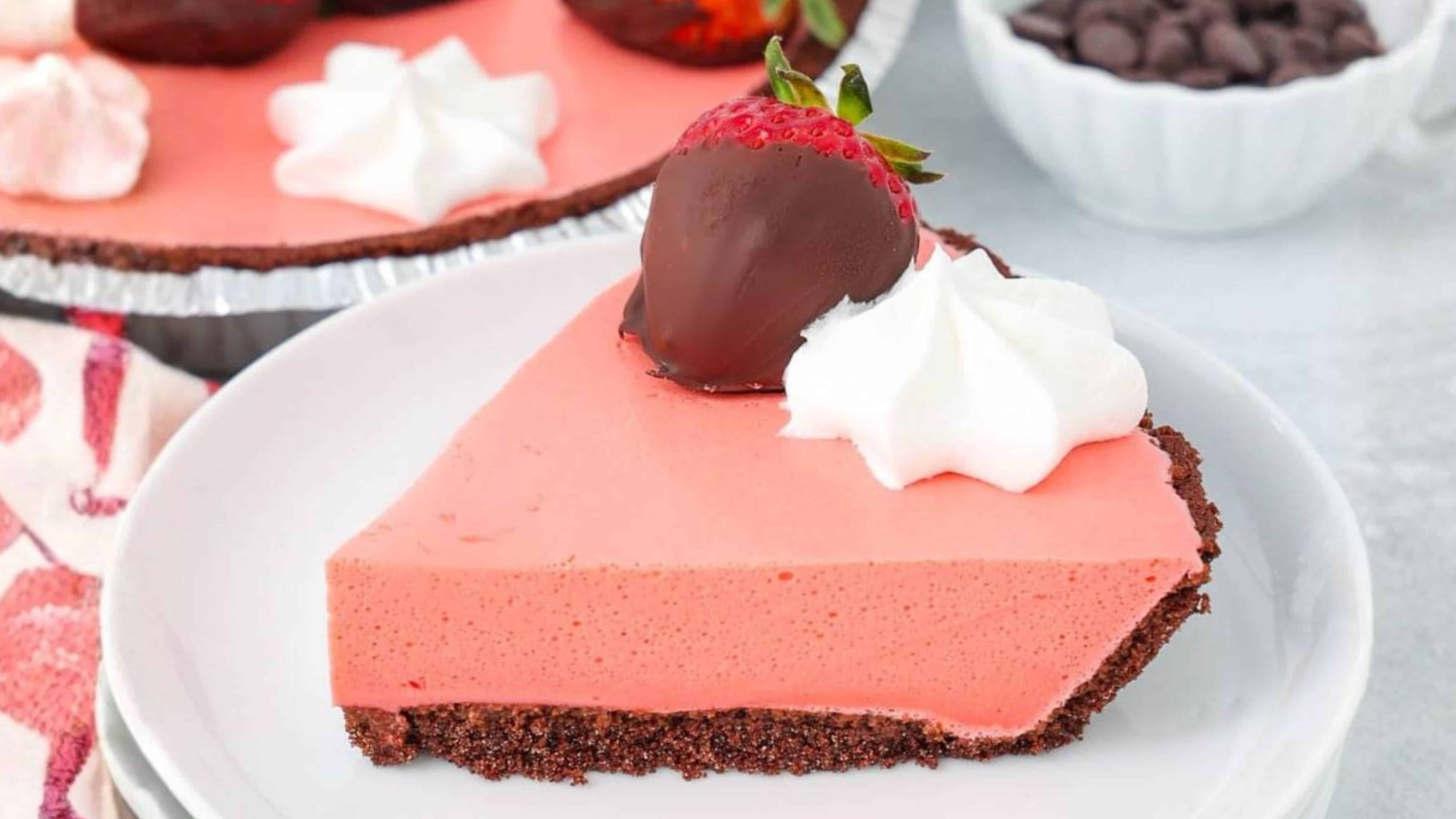 A slice of pink pie with a chocolate crust, topped with whipped cream and a chocolate-dipped strawberry.