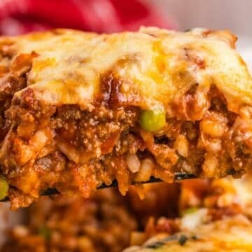 A close-up of a cheesy, baked lasagna slice with meat sauce, peas, and melted cheese being lifted from a pan.