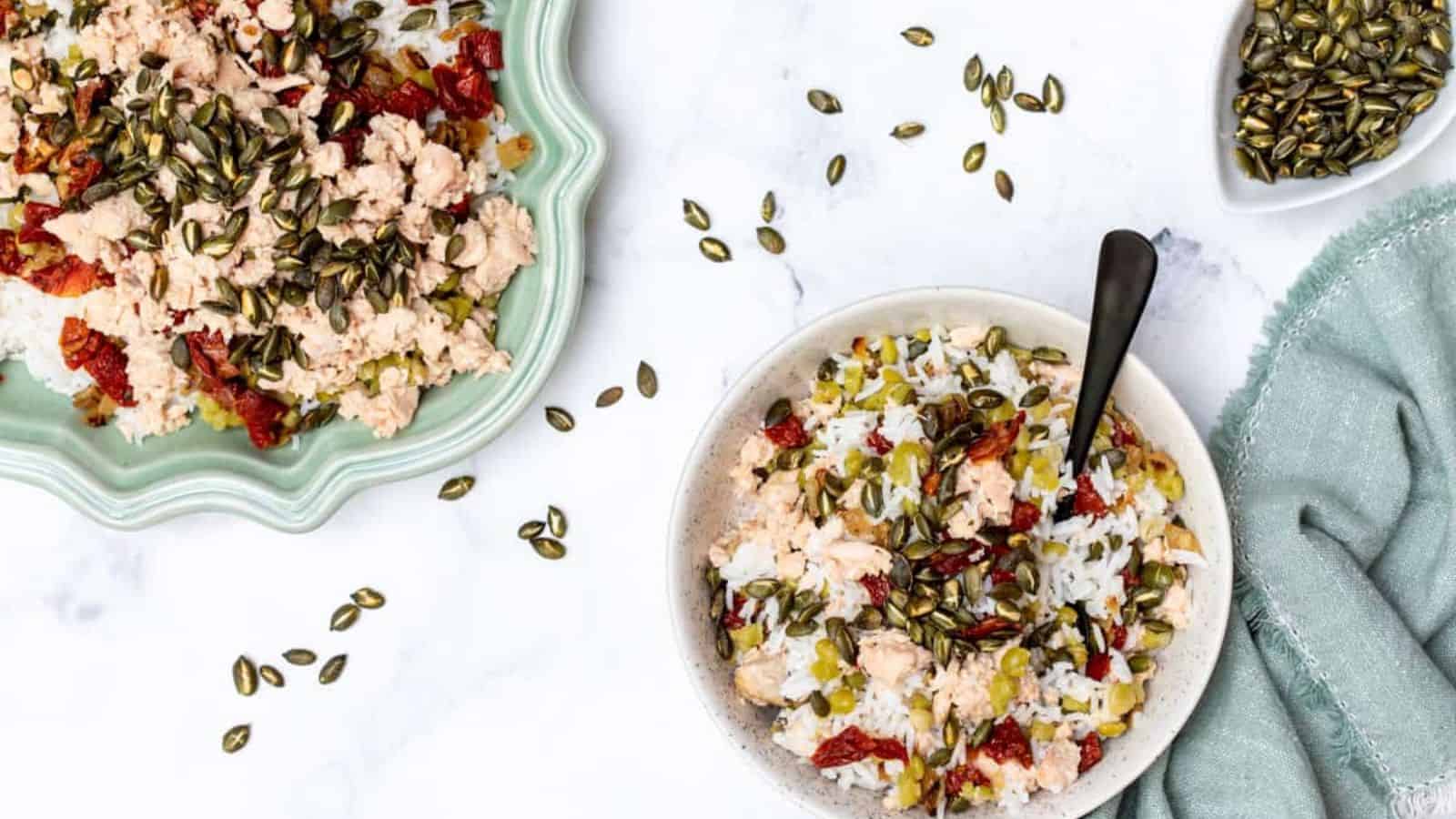A bowl of rice topped with ground turkey, sun-dried tomatoes, and pumpkin seeds beside a serving platter.