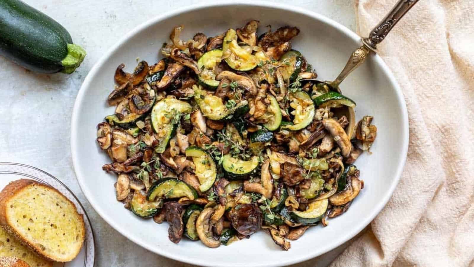 Saut&eacute;ed mushrooms and zucchini garnished with herbs in a white bowl, with bread and a zucchini nearby.