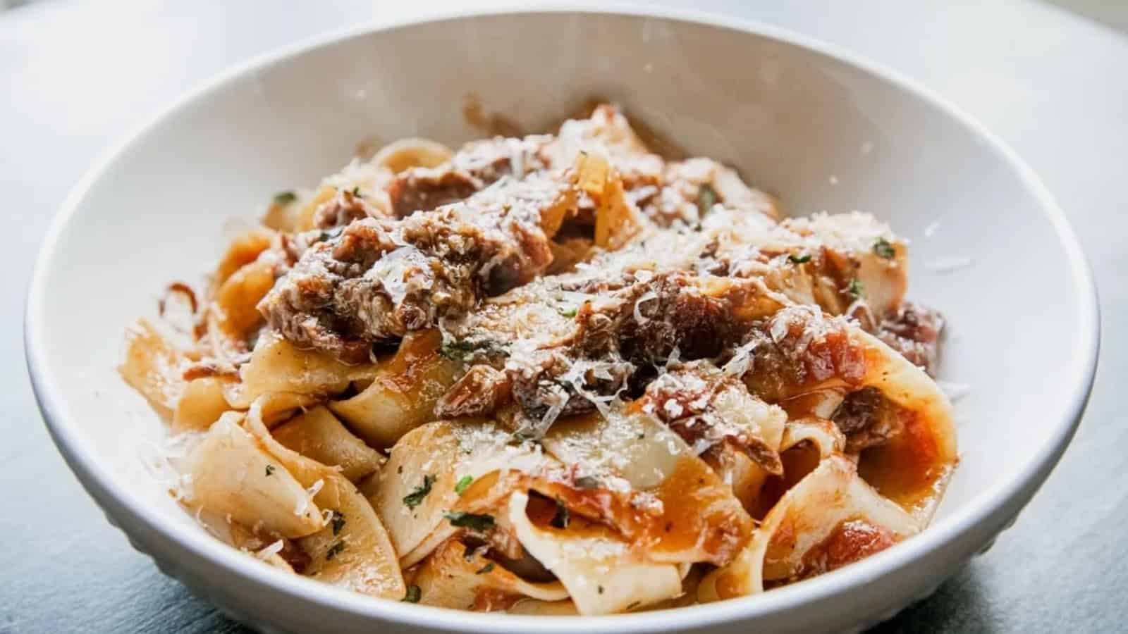 A white bowl filled with pappardelle pasta topped with a rich meat sauce and grated cheese.