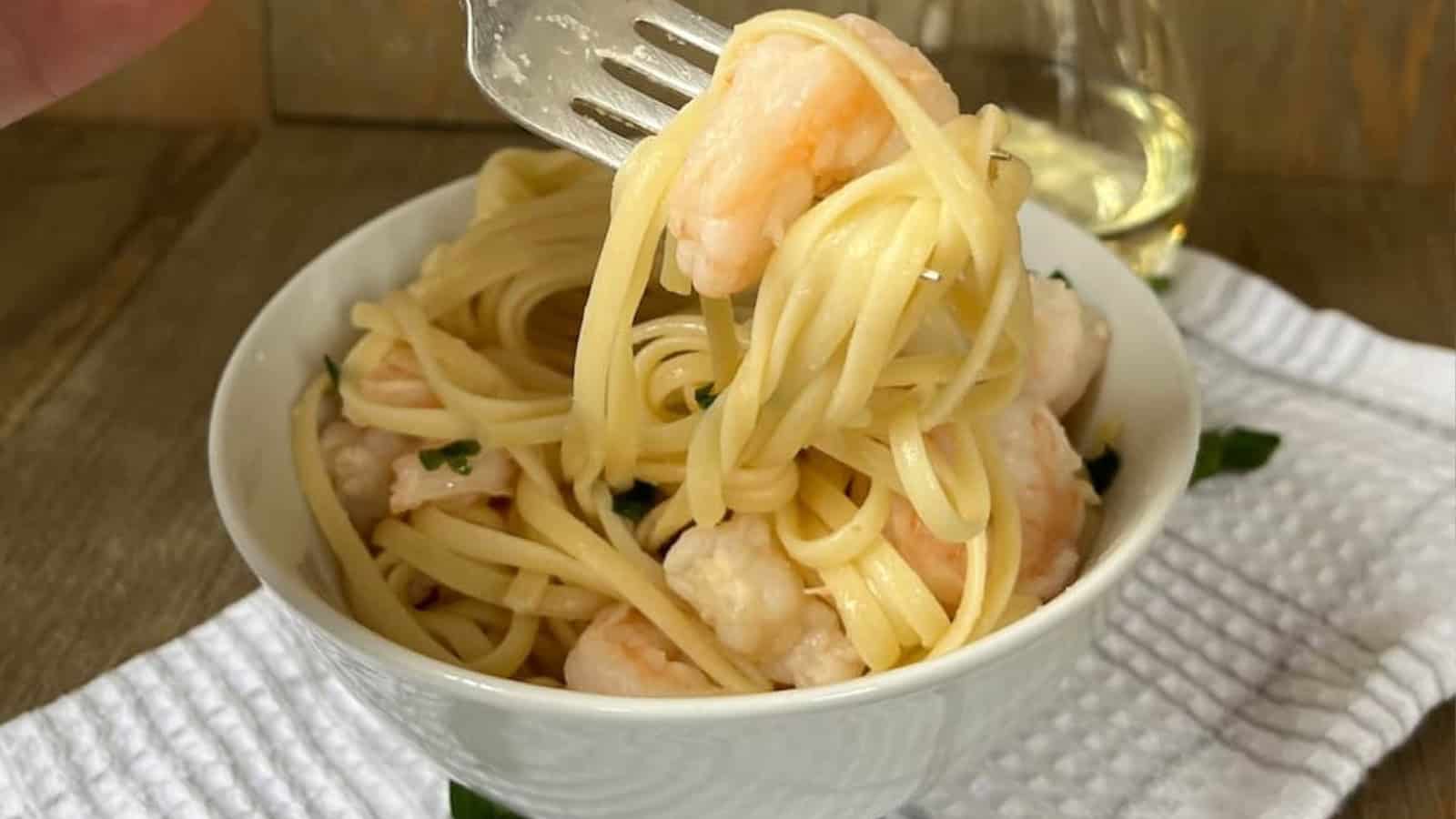 A hand holding a fork twirls spaghetti with shrimp in a white bowl, with a glass of white wine in the background on a wooden table.