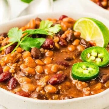 A bowl of chili with beans, ground meat, jalapeño slices, cilantro, and a lime wedge on top.