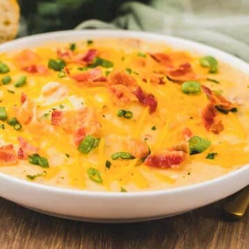 A bowl of creamy soup topped with shredded cheese, bacon bits, and chopped green onions on a wooden table.