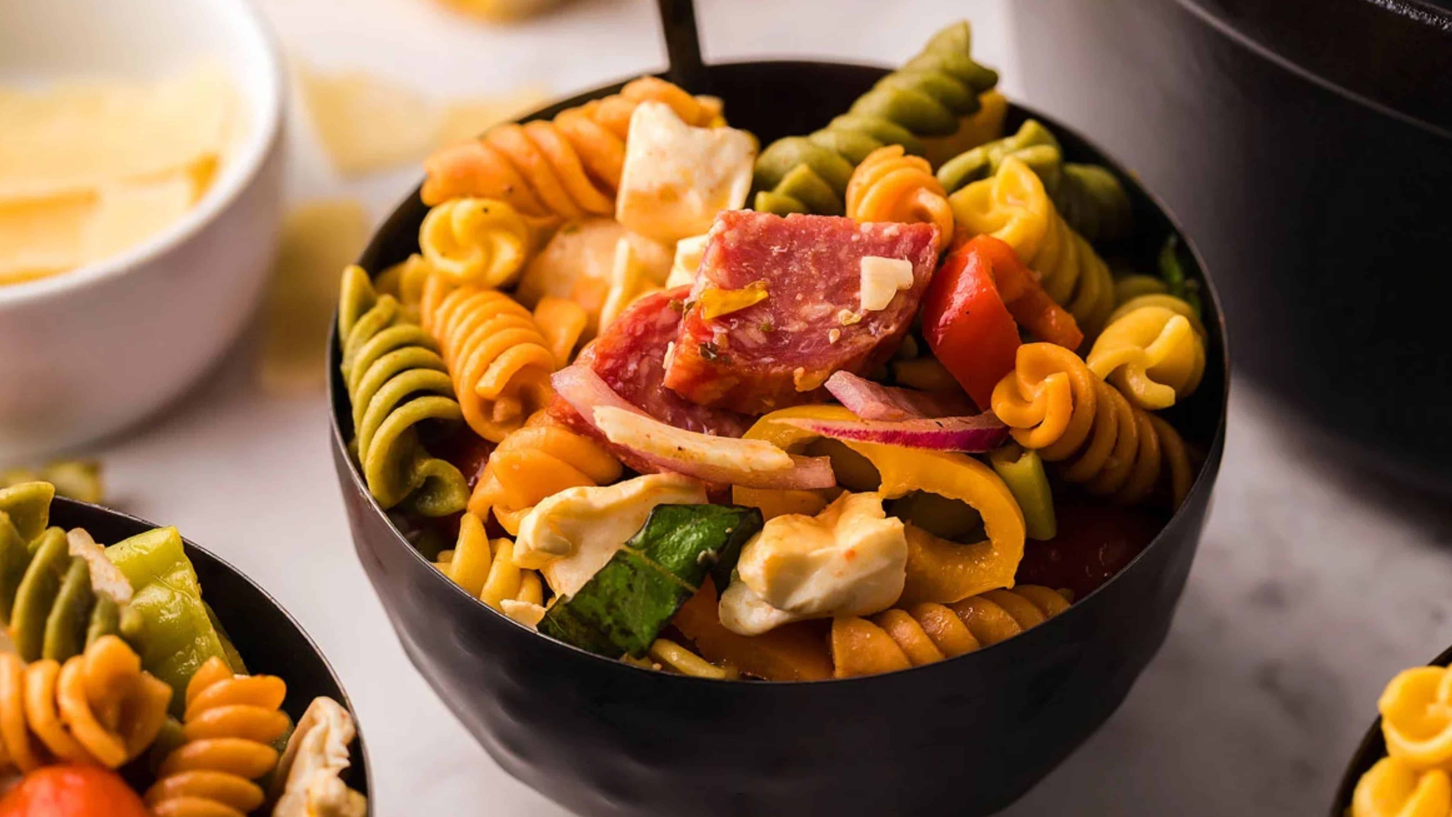 A bowl of colorful pasta salad with rotini, salami, mozzarella, tomatoes, onions, and peppers.