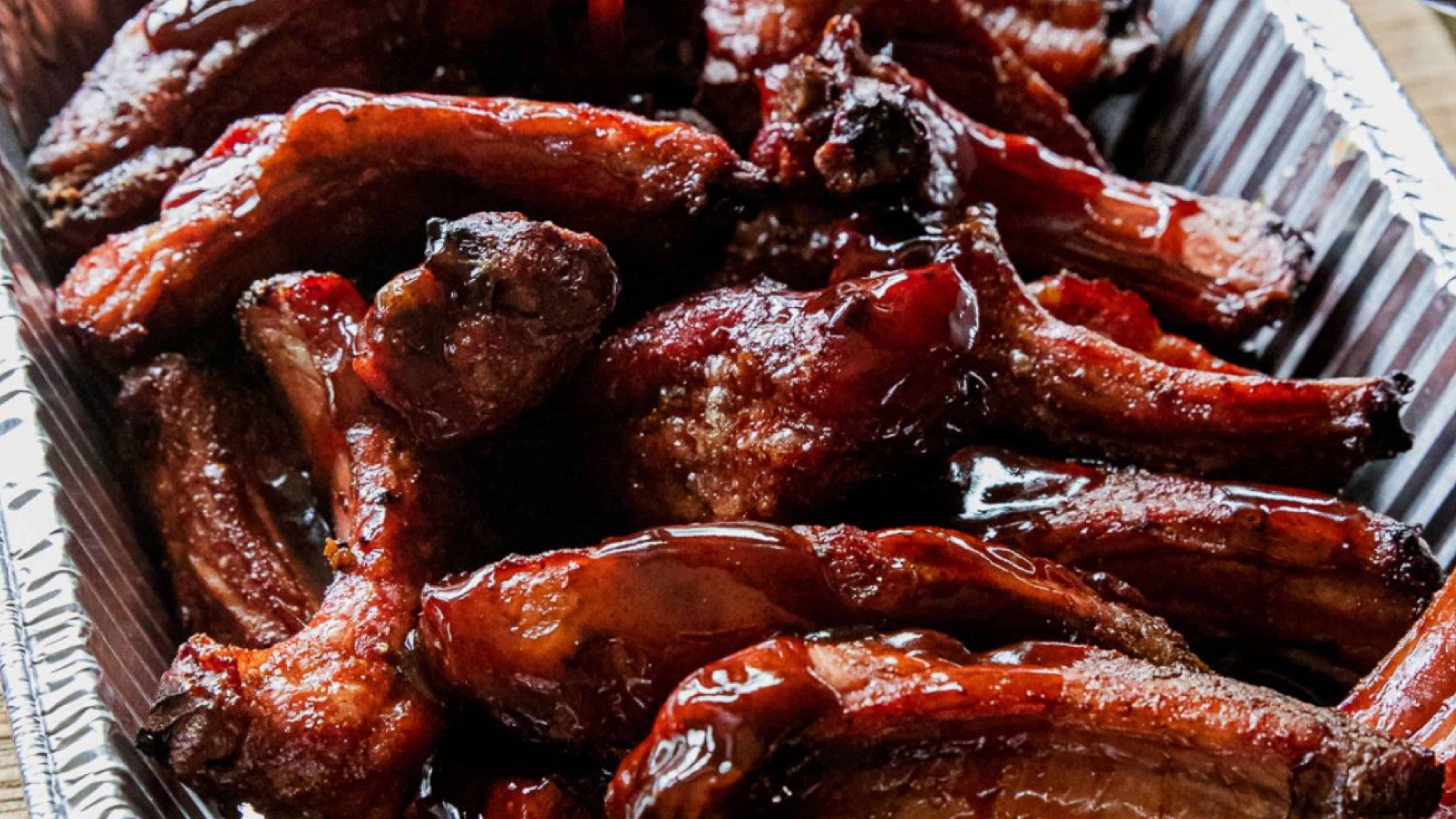 Glazed barbecued ribs piled in a foil tray, with a shiny, caramelized coating.