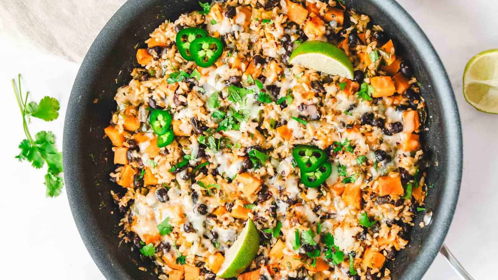 A skillet of rice, black beans, sweet potatoes, melted cheese, jalape&ntilde;os, cilantro, and lime wedges.