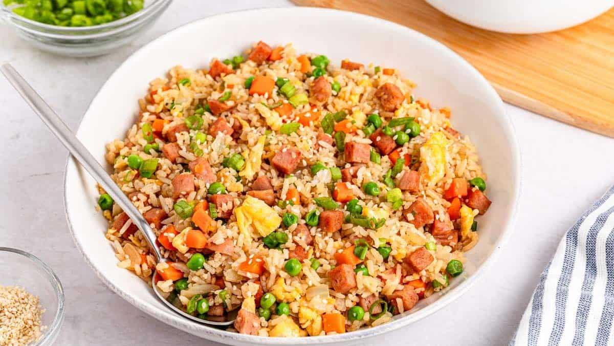 A bowl of fried rice with peas, carrots, scrambled eggs, and diced meat, served with a spoon.