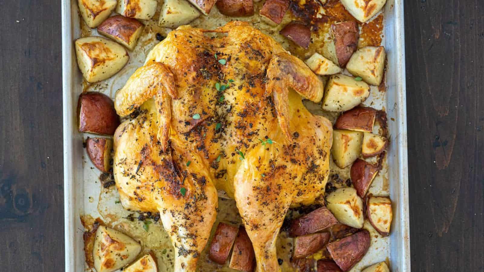 Roast chicken with herbs on a baking sheet, surrounded by golden-brown roasted potato pieces.