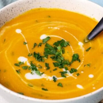 Bowl of creamy orange soup with a swirl of cream and chopped herbs, served with a black spoon.