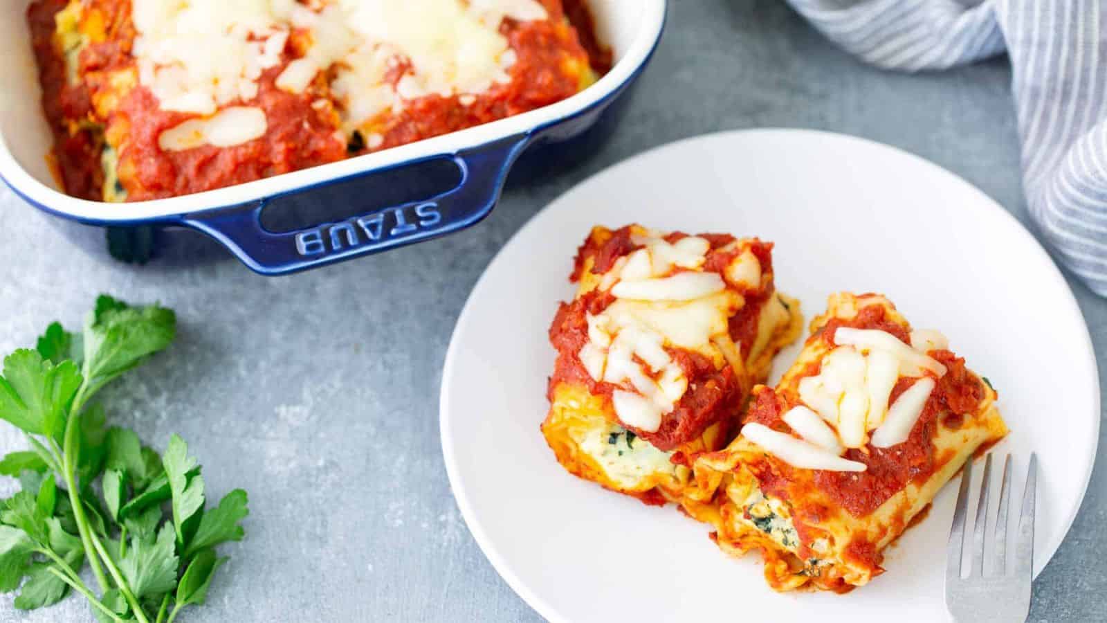 Two cheesy lasagna roll-ups on a white plate next to a dish of more lasagna; parsley on the side.