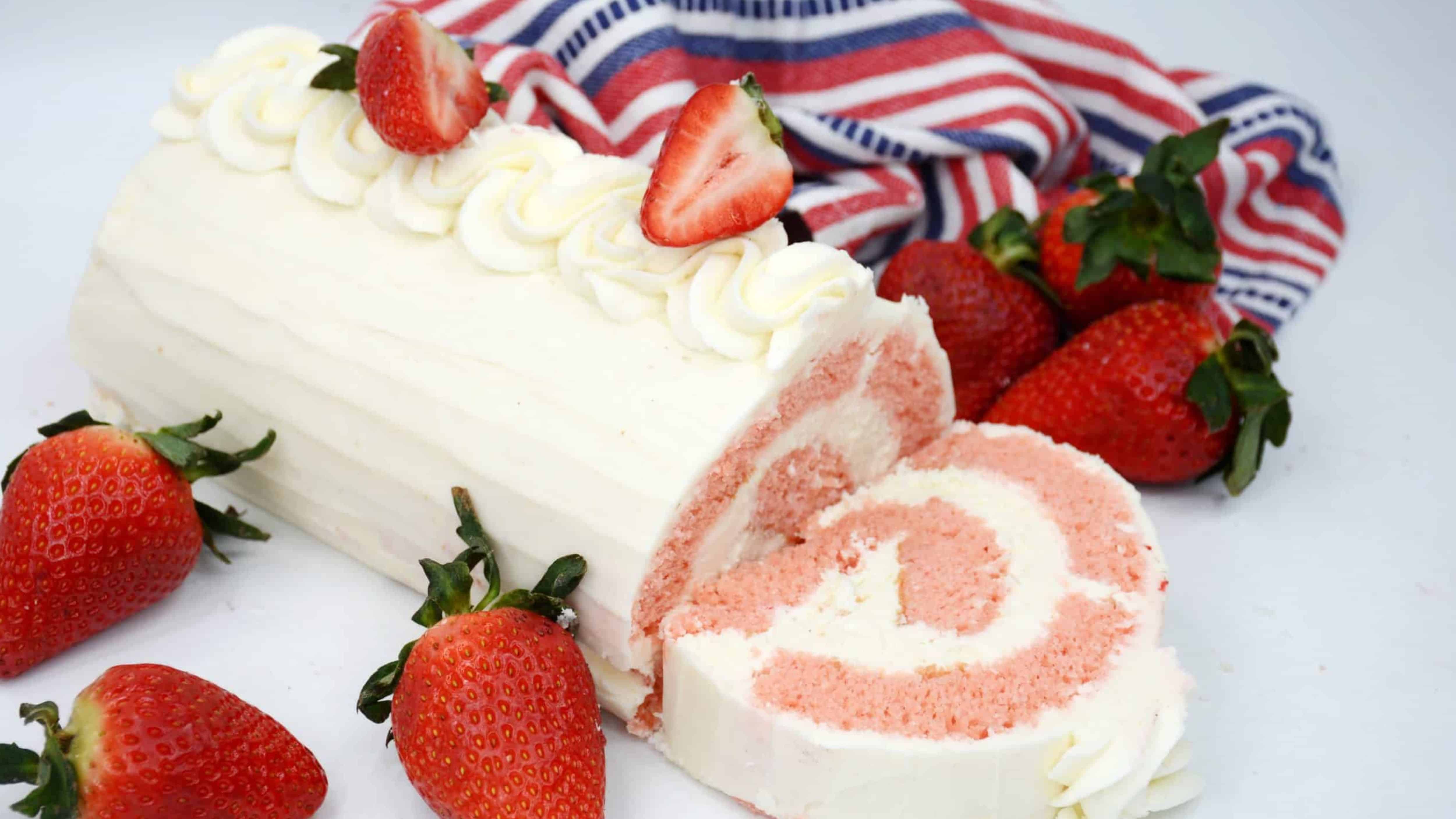 A strawberry cake roll with white frosting, topped with sliced strawberries, surrounded by whole strawberries.
