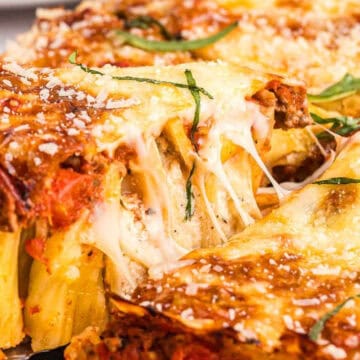 Cheesy baked pasta with tomato sauce, ground meat, and fresh basil being served from a casserole dish.