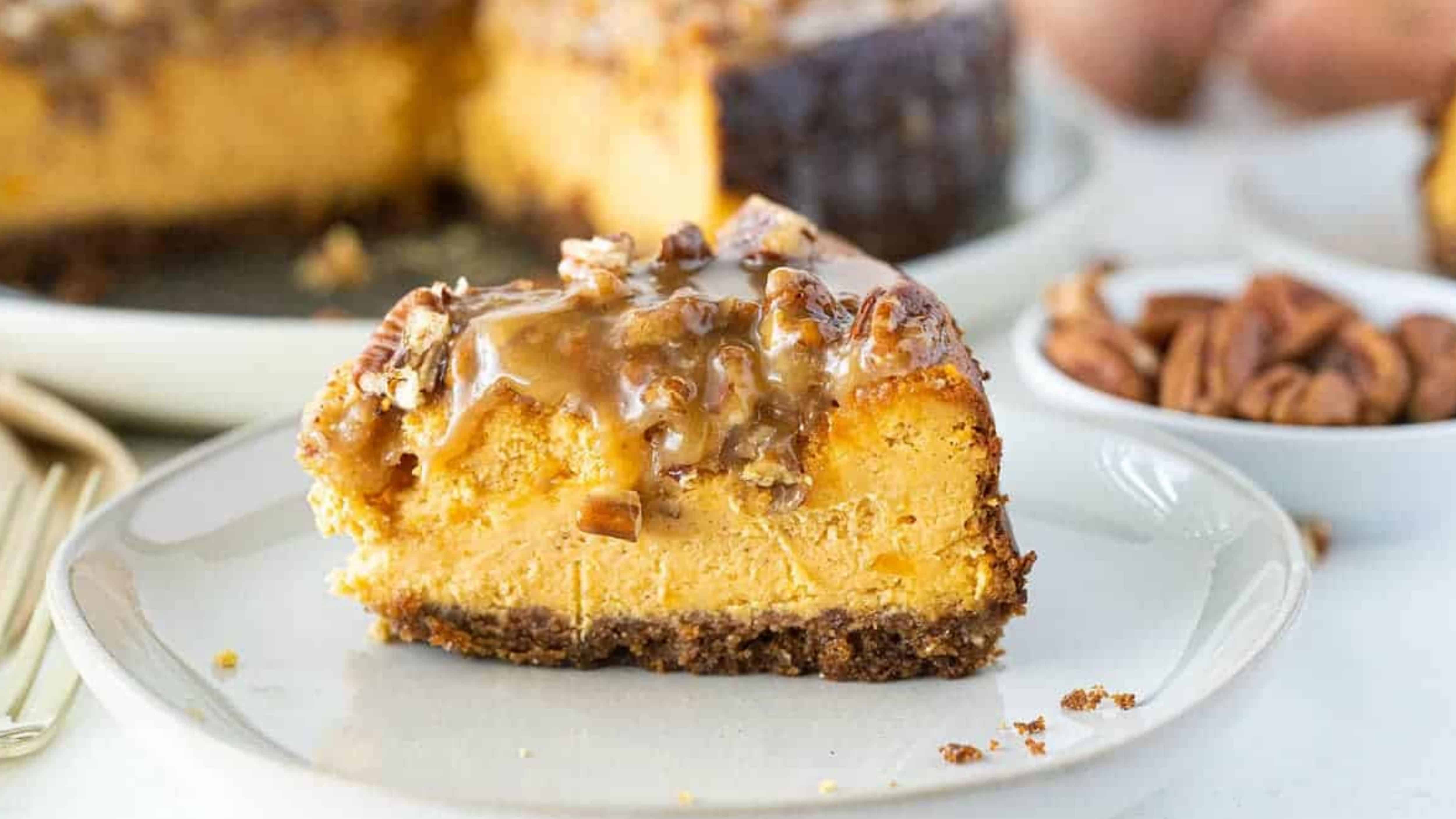 Slice of pumpkin cheesecake with pecan caramel topping on a plate, with nuts and more cheesecake in the background.