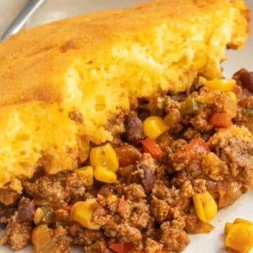 A slice of cornbread casserole with ground beef, corn, beans, and peppers on a white plate with a fork.