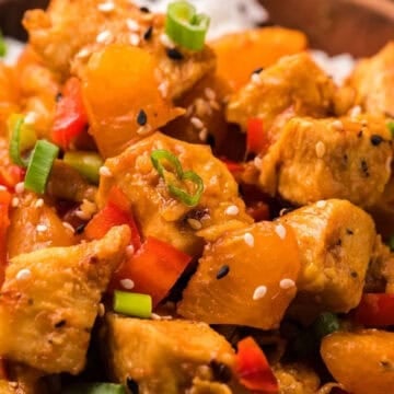 Close-up of orange chicken with bell peppers and green onions served over white rice, topped with sesame seeds.
