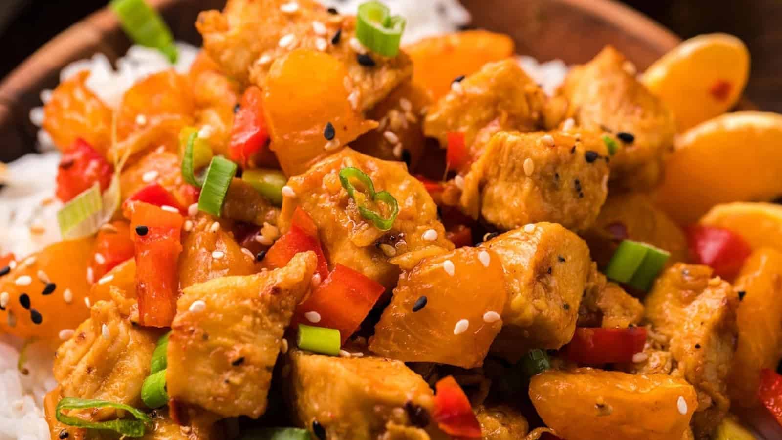 Close-up of orange chicken with bell peppers and green onions served over white rice, topped with sesame seeds.