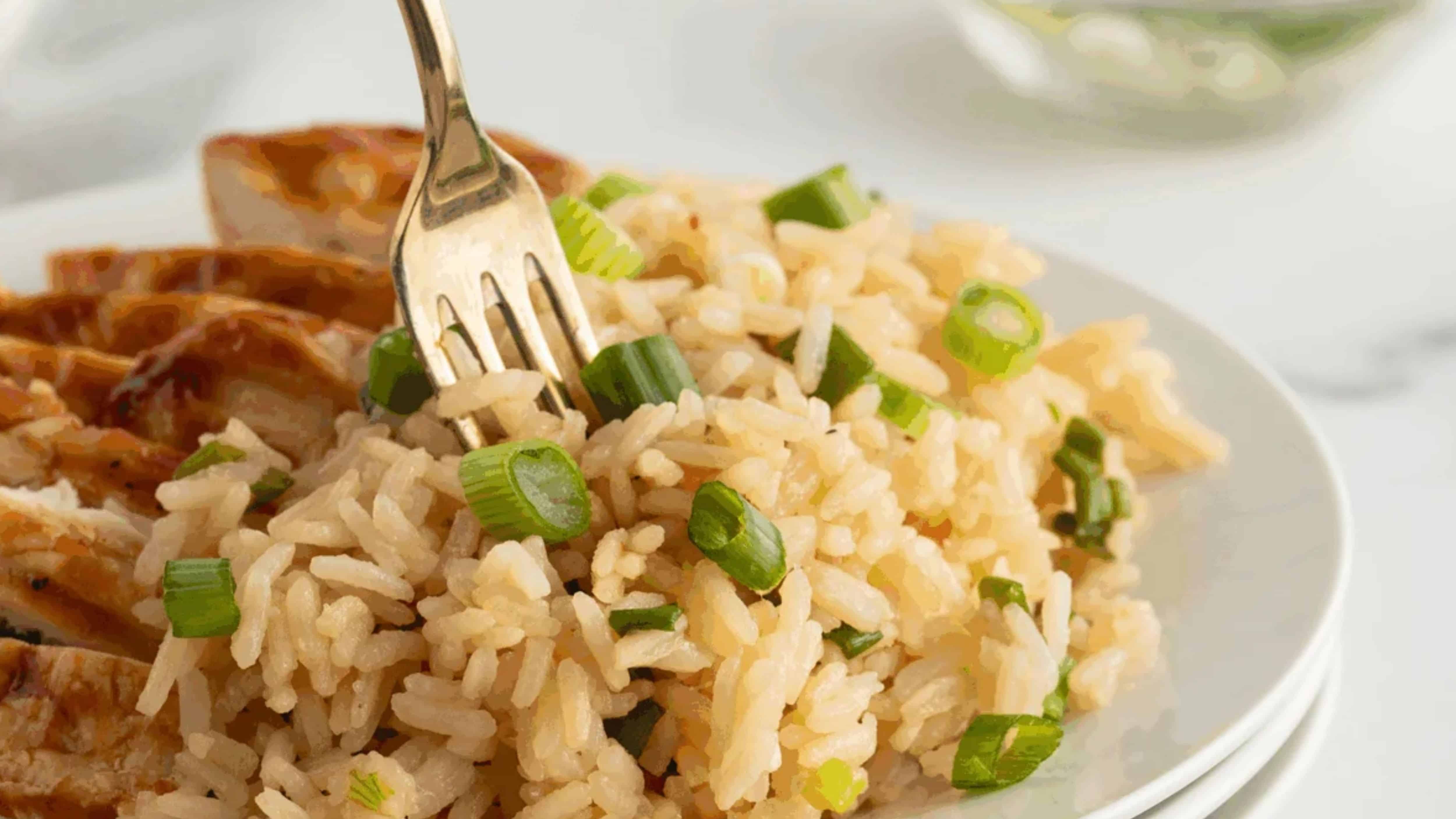 A fork in a plate of rice mixed with chopped green onions, with sliced chicken in the background.