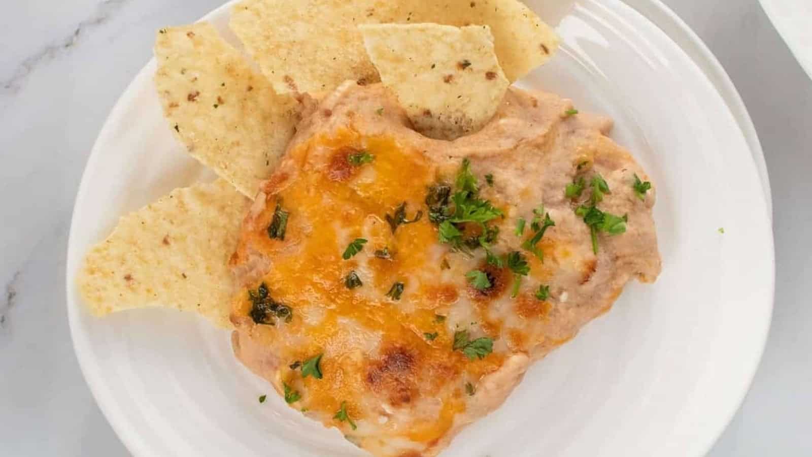 A plate of cheesy bean dip topped with parsley, served with tortilla chips on a white plate.