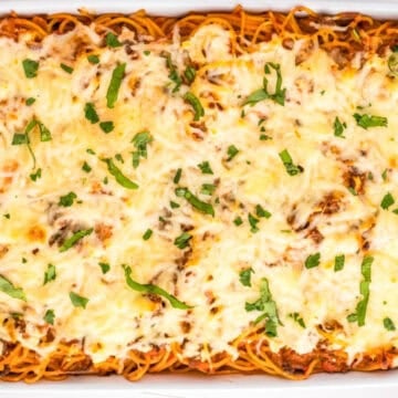Baked spaghetti casserole in a white dish, topped with melted cheese and chopped herbs.
