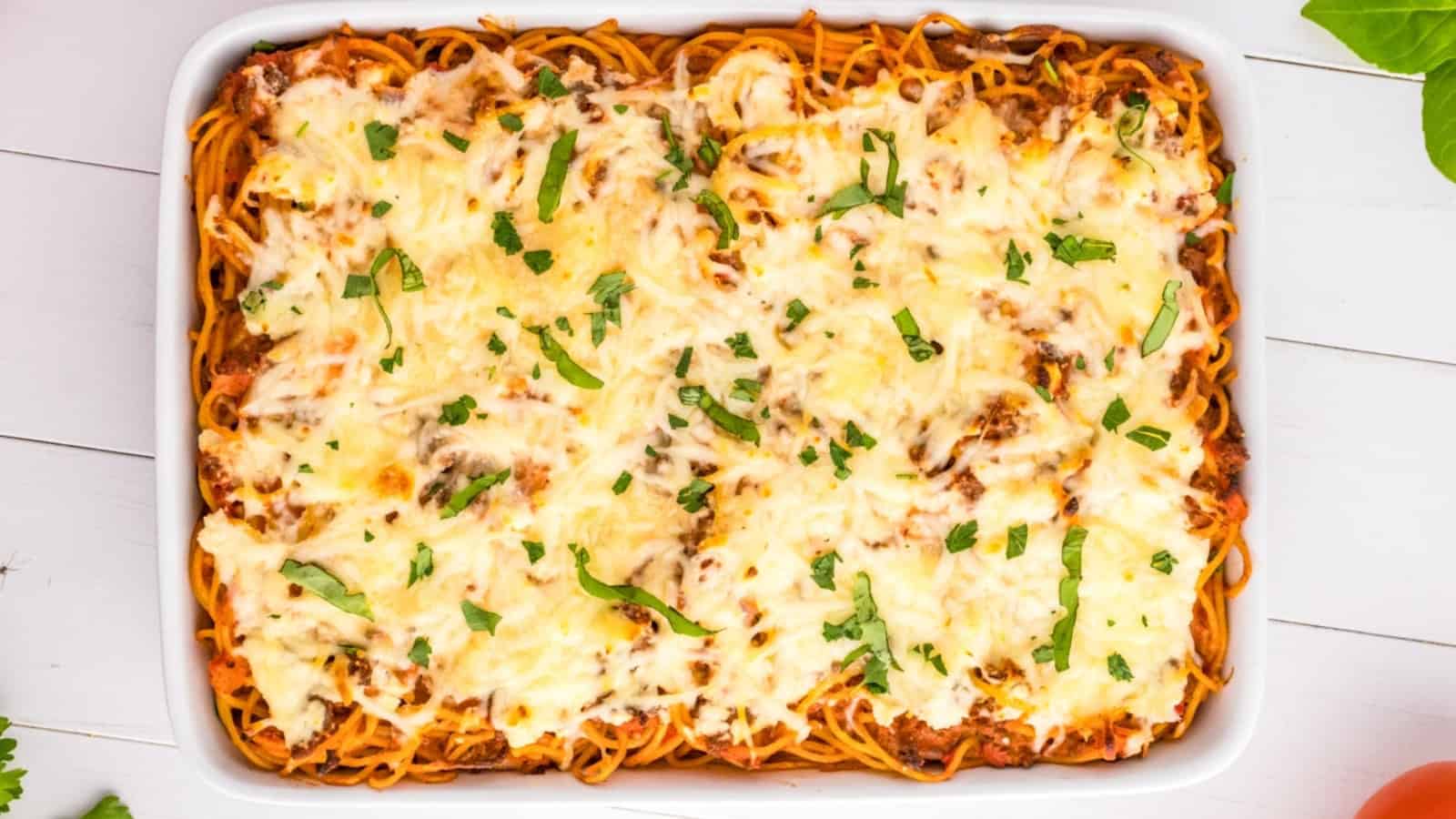 Baked spaghetti casserole in a white dish, topped with melted cheese and chopped herbs.