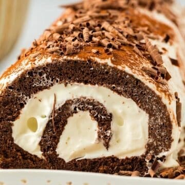 Chocolate Swiss roll cake with cream filling, topped with chocolate shavings, on a white plate.