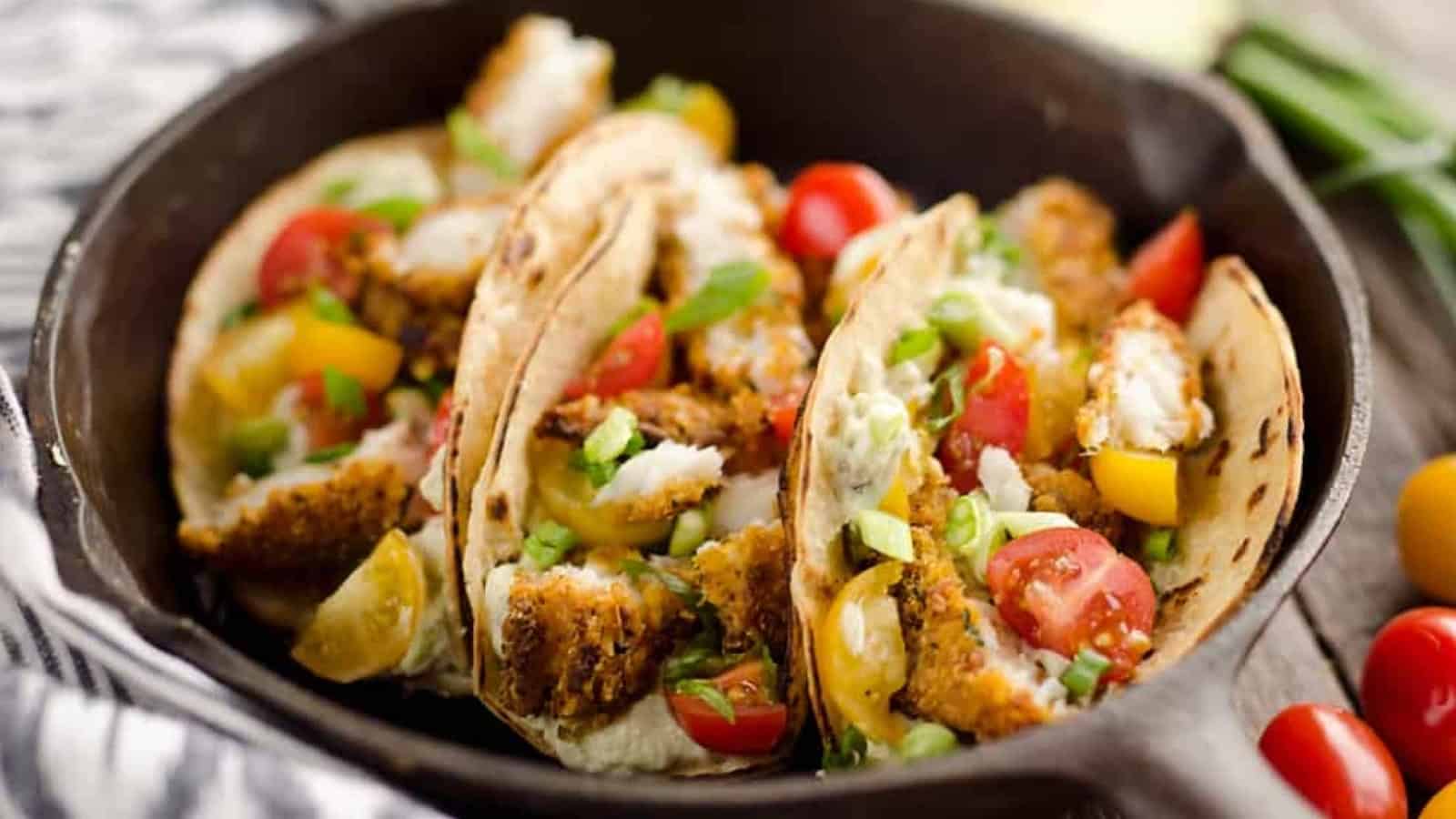 Three crispy fish tacos with diced tomatoes, bell peppers, and sauce in a black skillet.