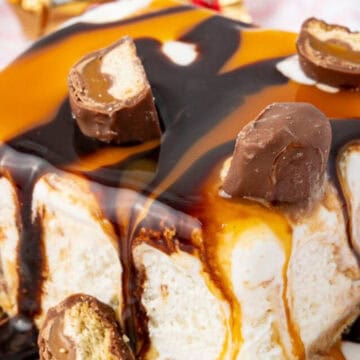 Ice cream cake topped with caramel, chocolate sauce, and pieces of Twix candy bars on a white plate.