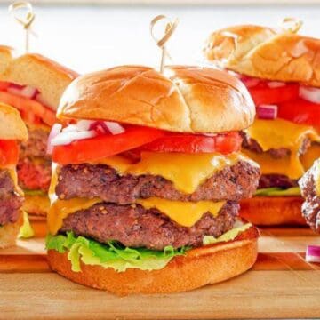 Four juicy cheeseburgers with lettuce, tomato, onions, and melted cheese on a wooden board.